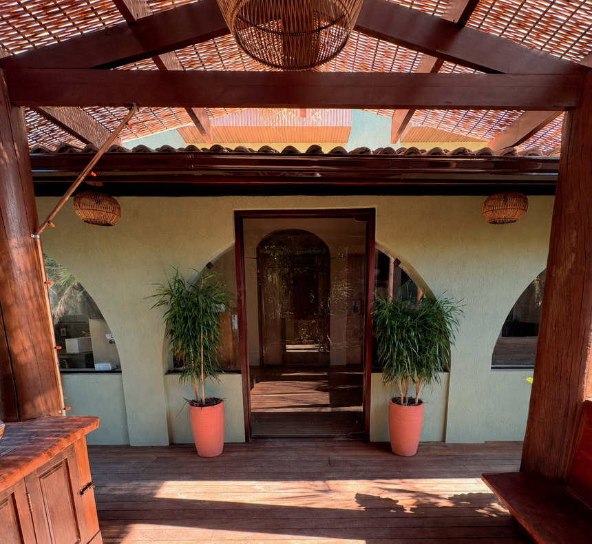 Entrada do Instituto Kerber: varanda de madeira com pergolado, luminárias de fibra, vasos com plantas e portal de vidro entre arcos